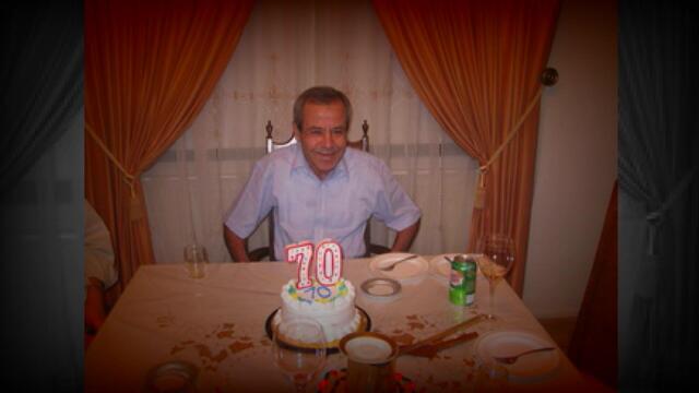 Chafic "Steve" Ezzeddine sits at a table with a birthday cake with candles in the shape of the number "70." 